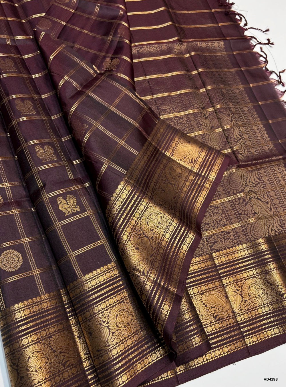 The Dark Coffee Brown Checkered Kanchi Border Soft Silk Saree is a refined handwoven creation that radiates earthy elegance and cultural grandeur. The deep coffee brown shade conveys warmth, devotion, and timeless sophistication, while the checkered weave adds a touch of rustic charm and heritage appeal. The temple‑inspired Kanchi border enriched with pure gold zari elevates its grandeur, making this saree perfect for weddings, temple ceremonies, and festive occasions. Its lustrous drape and coordinated blo