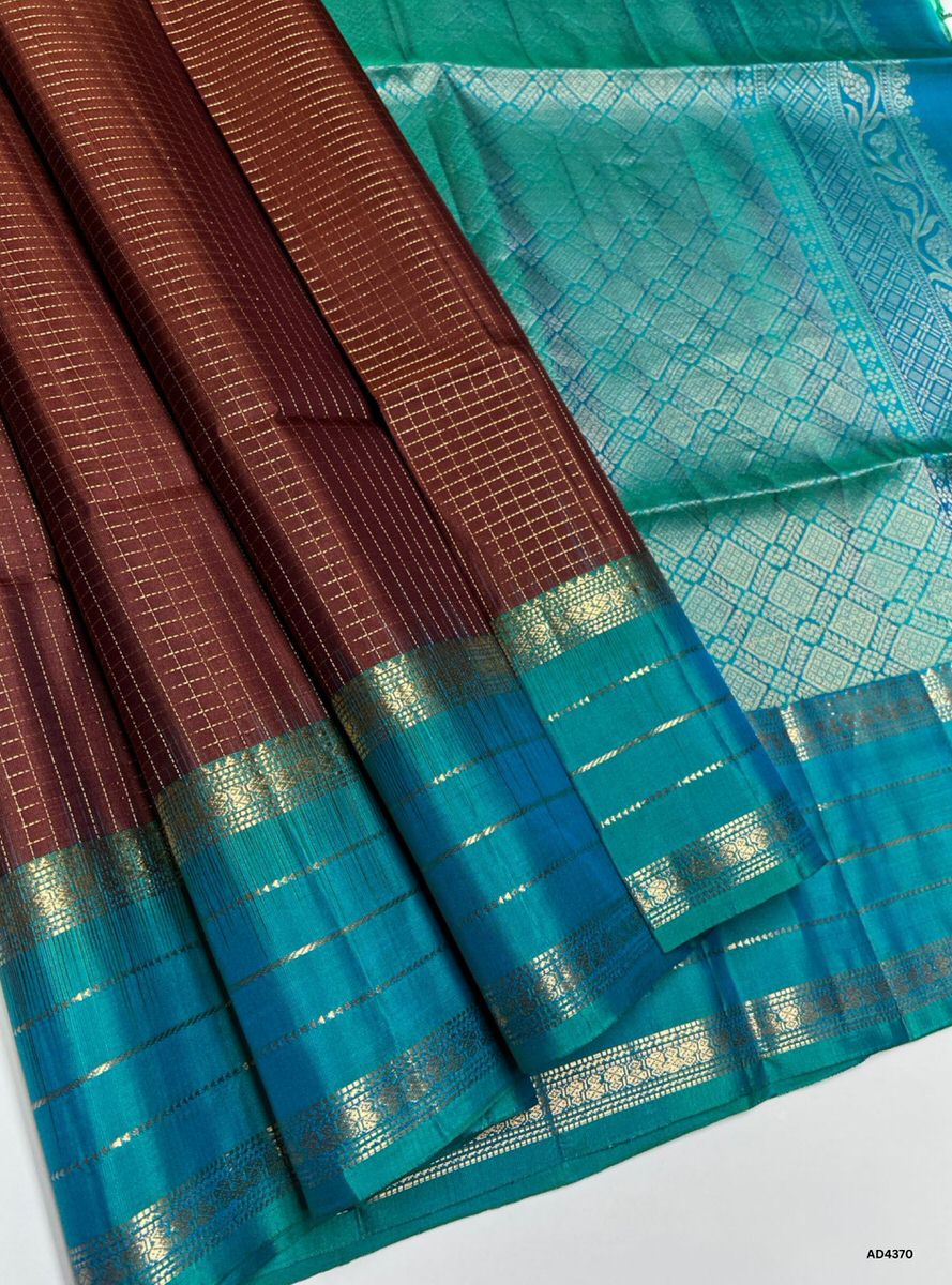 The Brown & Rama Blue Checkered Retta Pet Soft Silk Saree is a refined handwoven creation that radiates cultural richness and festive charm. The brown shade conveys devotion, warmth, and heritage depth, while the Rama blue checks symbolize prosperity, vibrance, and timeless elegance. Enhanced with pure gold zari borders and a lustrous drape, this saree is perfect for weddings, temple ceremonies, and celebratory occasions. A timeless masterpiece, it blends earthy tones with jewel‑tone artistry in the traditi