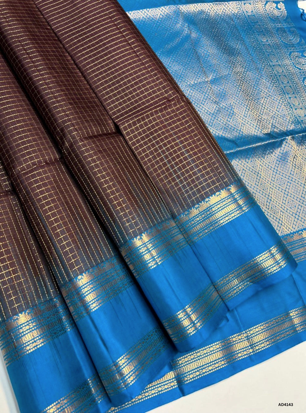The Coffee Brown & Rama Blue Checkered Retta Pet Soft Silk Saree is a striking handwoven creation that radiates festive elegance and cultural richness. The coffee brown shade conveys warmth, tradition, and earthy depth, while the rama blue adds celebratory brilliance and youthful vibrance. The checkered weave across the body enhances its visual appeal, while the Retta Pet temple‑inspired double line border enriches the saree with timeless grandeur. Enhanced with pure gold zari and a lustrous drape, this sar