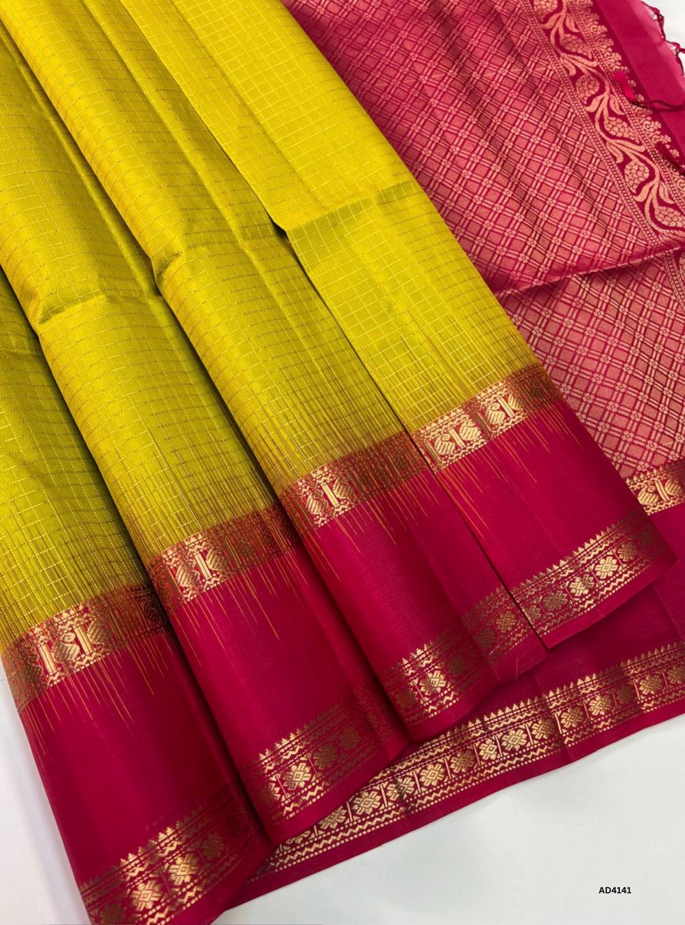 The Golden Yellow & Rani Pink Checkered Retta Pet Soft Silk Saree is a radiant handwoven creation that celebrates festive grandeur and cultural heritage. The golden yellow shade conveys prosperity, auspiciousness, and celebratory brilliance, while the rani pink adds vibrance, joy, and regal charm. The checkered weave across the body enhances its visual appeal, while the Retta Pet temple‑inspired double line border enriches the saree with timeless artistry. Enhanced with pure gold zari and a lustrous drape,