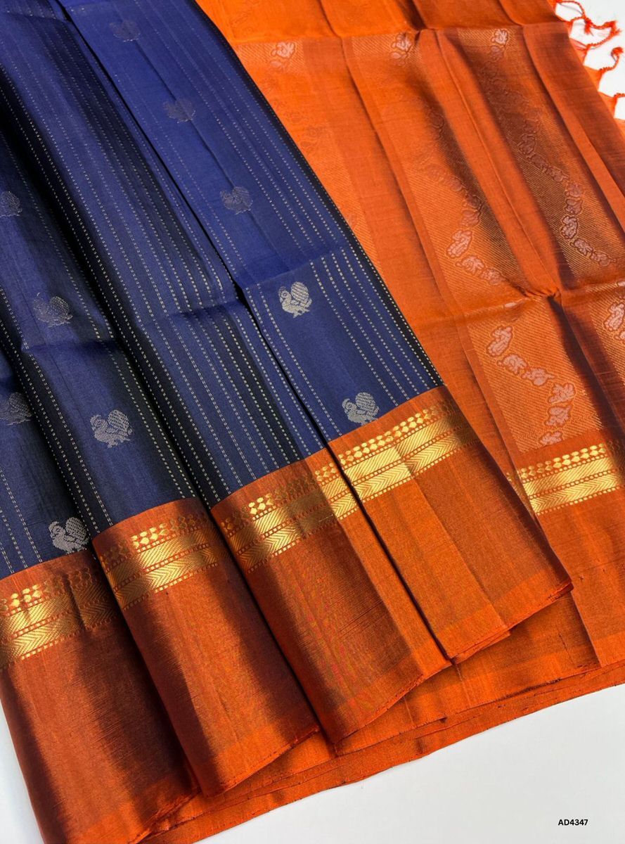The Navy Blue & Rust Vairoosi X Kanchi Border Soft Silk Saree is a majestic handwoven creation that radiates festive grandeur and cultural richness. The navy blue shade symbolizes devotion, serenity, and timeless elegance, while the rust contrast conveys warmth, heritage depth, and celebratory charm. The unique Vairoosi X Kanchi border highlights dual weaving traditions, making this saree a collector’s piece. Enhanced with pure gold zari and a lustrous drape, it is perfect for weddings, temple ceremonies, a