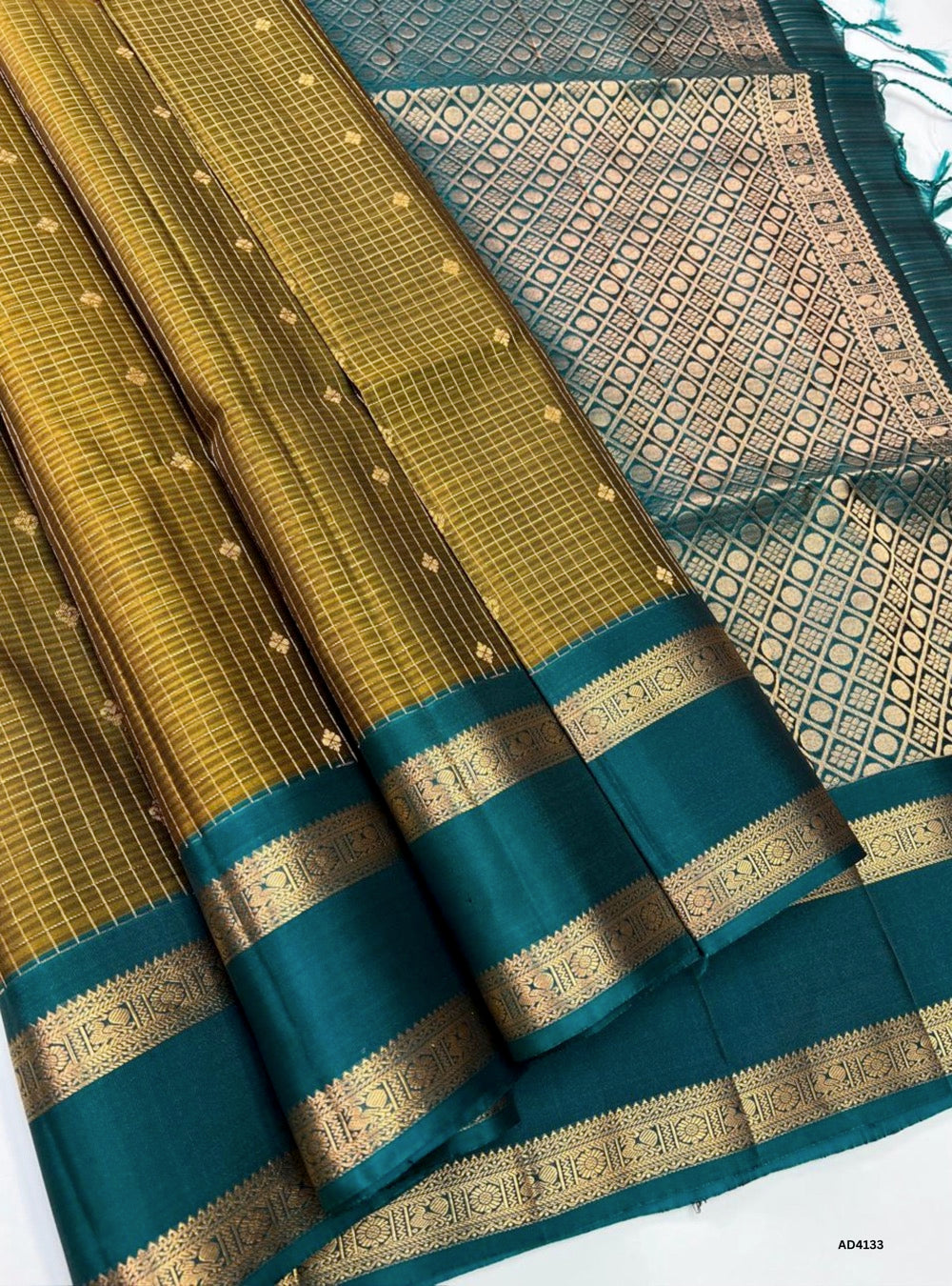 The Olive Green & Rama Green Checkered Retta Pet Soft Silk Saree is a heritage handwoven creation that radiates festive charm and cultural richness. The olive green shade conveys serenity, tradition, and earthy elegance, while the rama green adds celebratory brilliance and youthful vibrance. The checkered weave across the body enhances its visual appeal, while the Retta Pet temple‑inspired double line border enriches the saree with timeless grandeur. Enhanced with pure gold zari and a lustrous drape, this s
