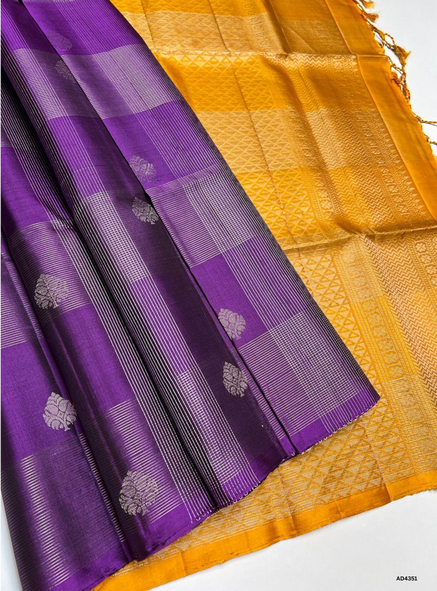 The Purple & Mustard Yellow Checkered Soft Silk Saree is a striking handwoven creation that radiates festive brilliance and cultural richness. The regal purple shade symbolizes devotion, sophistication, and heritage depth, while the mustard yellow checks add vibrance, auspiciousness, and celebratory warmth. Enhanced with pure gold zari borders and a lustrous drape, this saree is perfect for weddings, temple ceremonies, and festive occasions. A timeless masterpiece, it blends jewel‑tone grandeur with checker