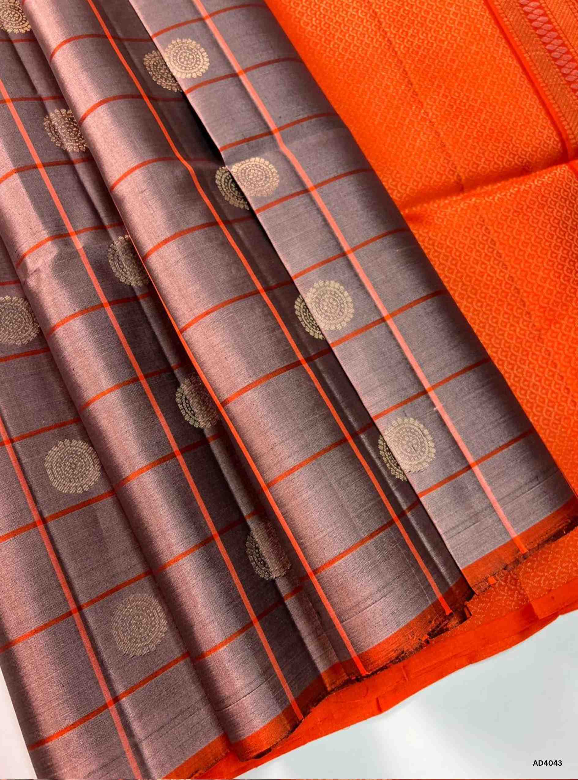 The Stone Grey & Orange Checkered Soft Silk Saree is a striking handwoven creation that radiates festive charm and cultural richness. The subtle stone grey shade symbolizes calm elegance, while the vibrant orange adds auspicious brilliance and celebratory warmth. The checkered weave, enhanced with delicate motifs, creates a rhythmic visual appeal, while zari highlights on the border and pallu lend a shimmering finish. Perfect for weddings, temple ceremonies, and cultural gatherings, this saree embodies Sout