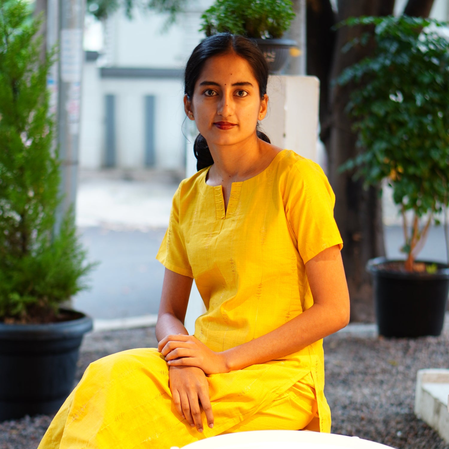 Person wearing a yellow outfit standing outdoors with plants and a white table in the background