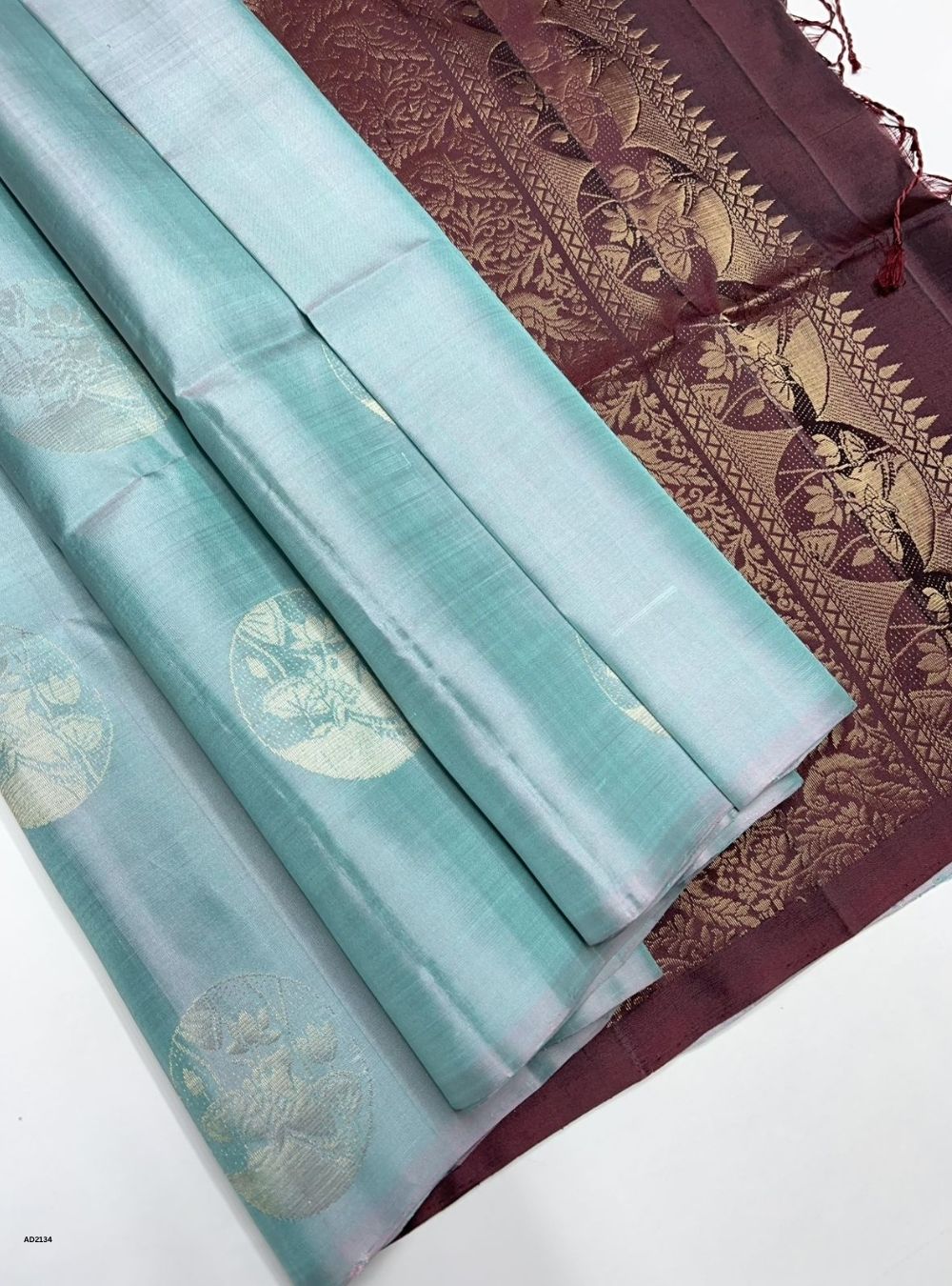 Two sarees, one light blue with gold patterns and the other brown with intricate designs, on a white background.