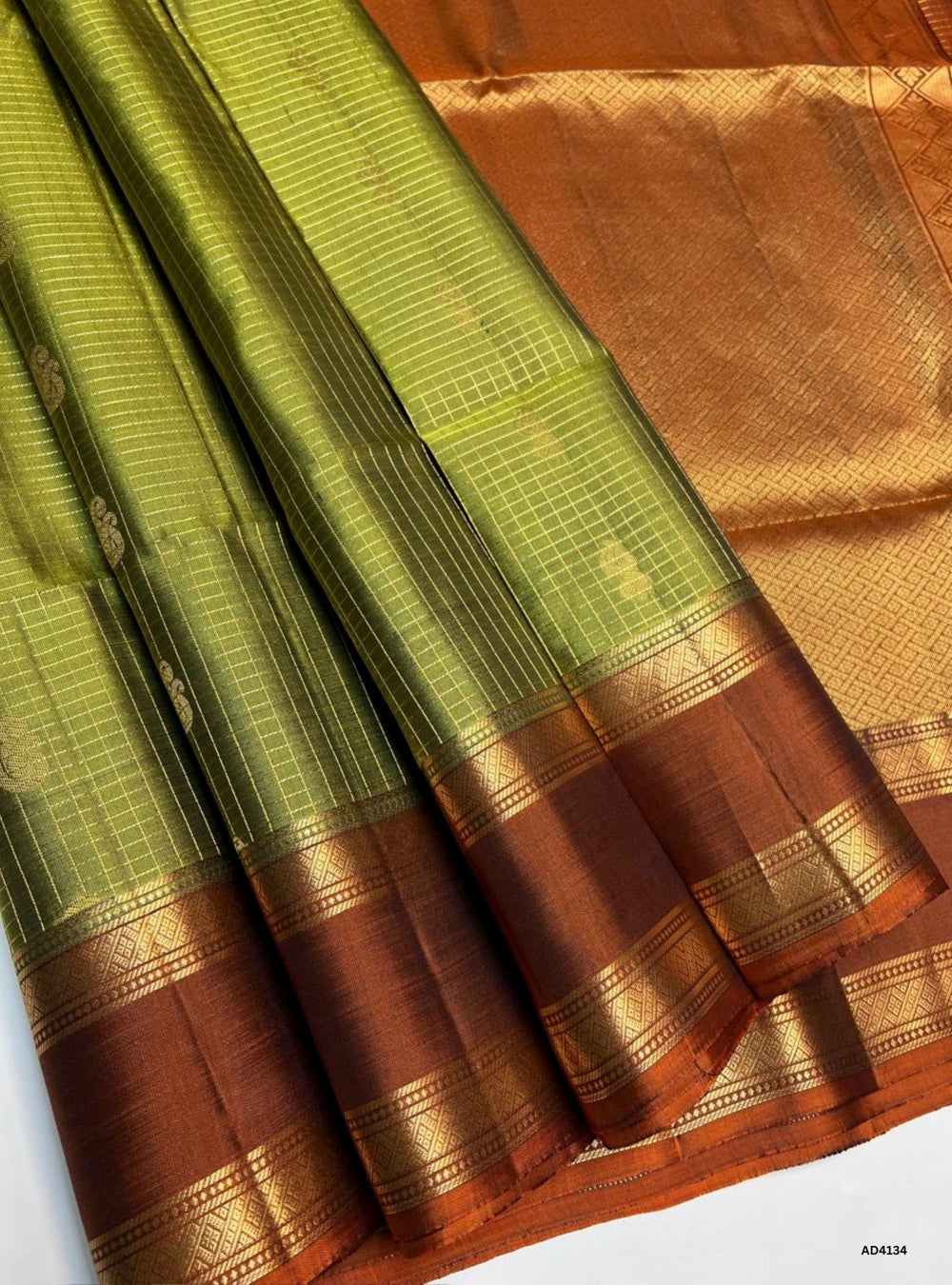 The Pea Green & Brown Checkered Retta Pet Soft Silk Saree is a charming handwoven creation that radiates festive elegance and cultural richness. The pea green shade conveys freshness, prosperity, and youthful vibrance, while the coffee brown adds warmth, depth, and traditional grandeur. The checkered weave across the body enhances its visual appeal, while the Retta Pet temple‑inspired double line border enriches the saree with heritage artistry. Enhanced with pure gold zari and a lustrous drape, this saree 